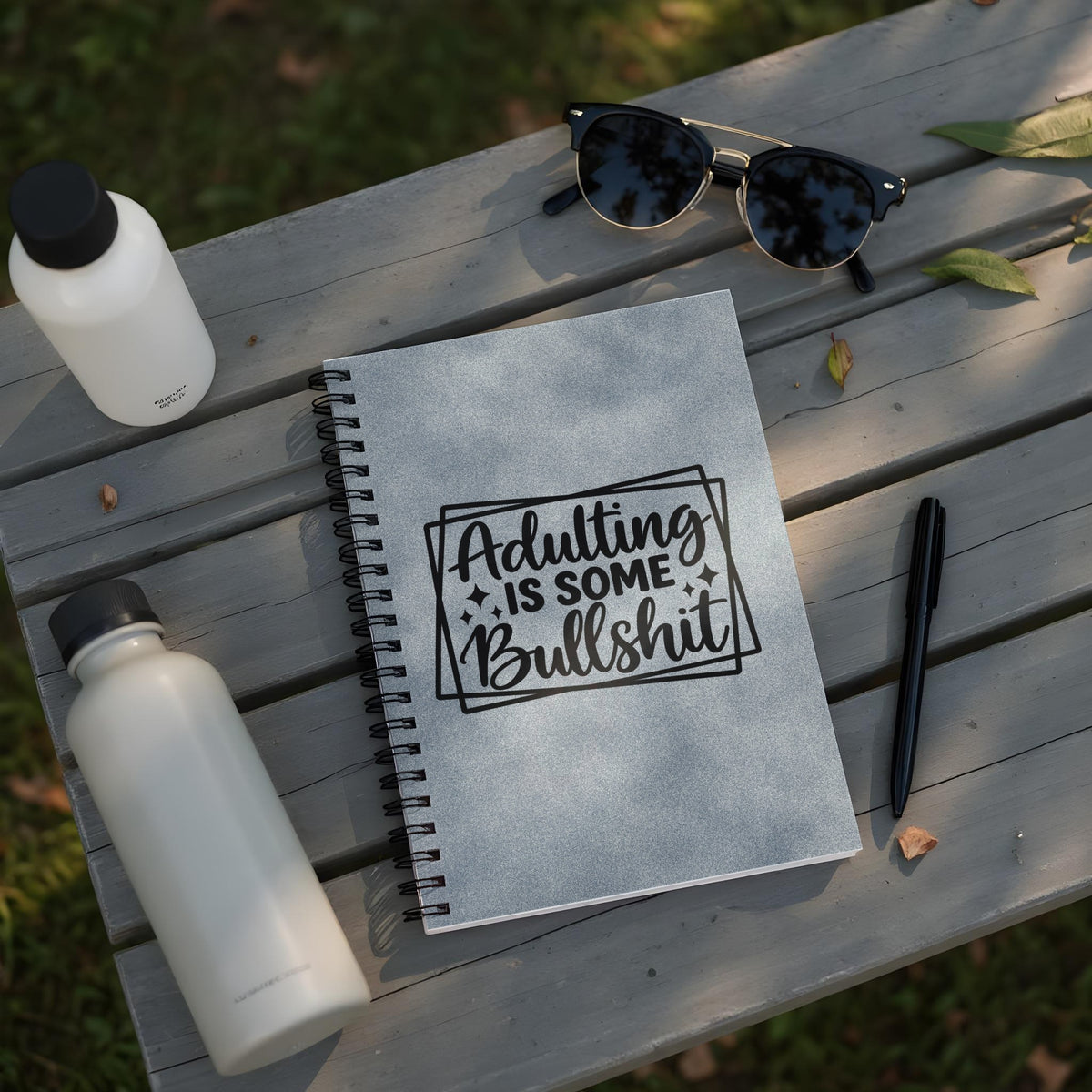 Spiral notebook with "Adulting is Some Bullshit" text on a wooden surface with water bottles.